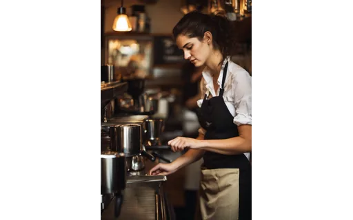 A barista in her twenties behind a busy espresso machine in a small cafe, steam rising, hands working the portafilter, warm interior light